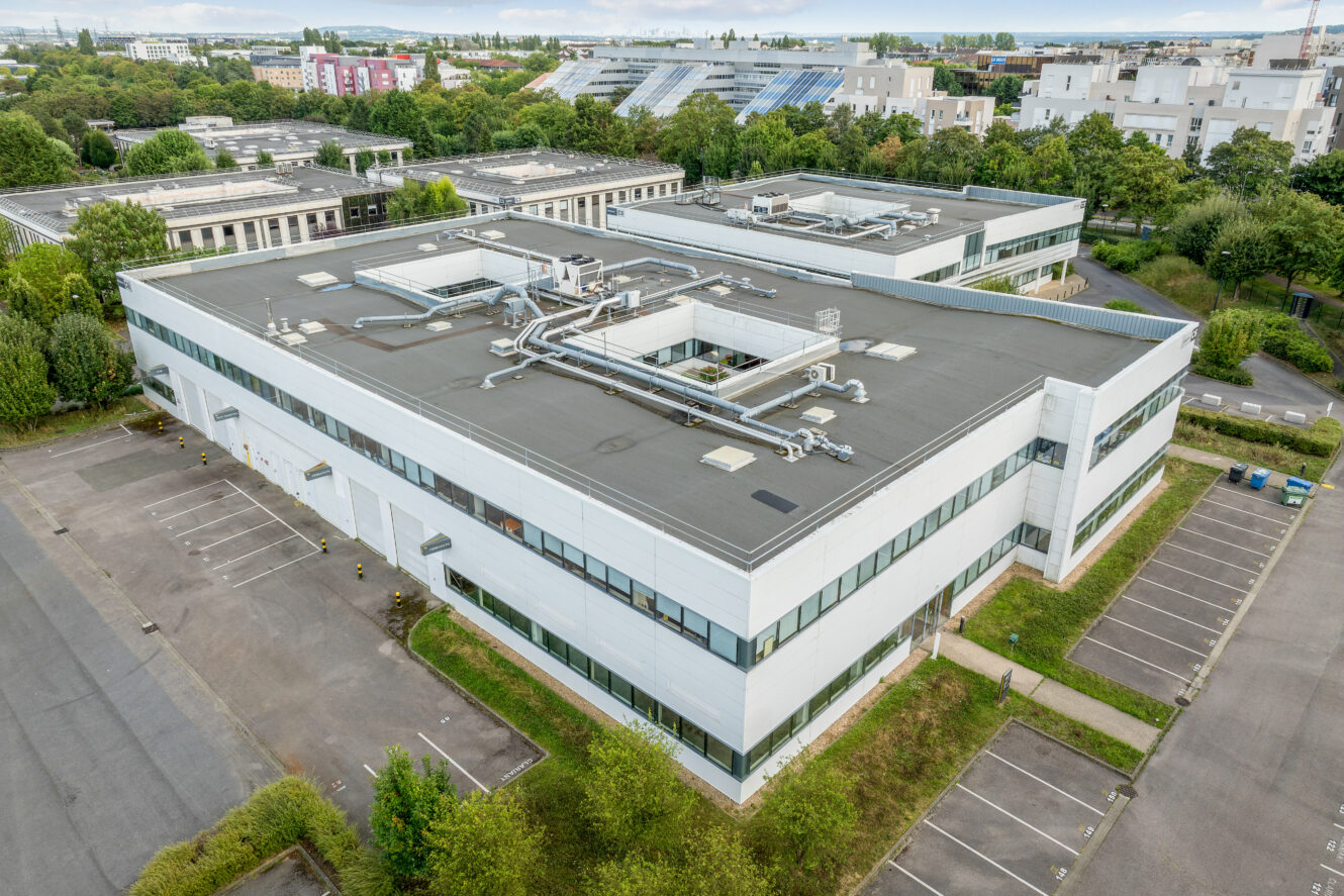 Un grand bâtiment industriel ou de bureaux moderne avec des systèmes de ventilation sur le toit, entouré de places de parking vides et de verdure.