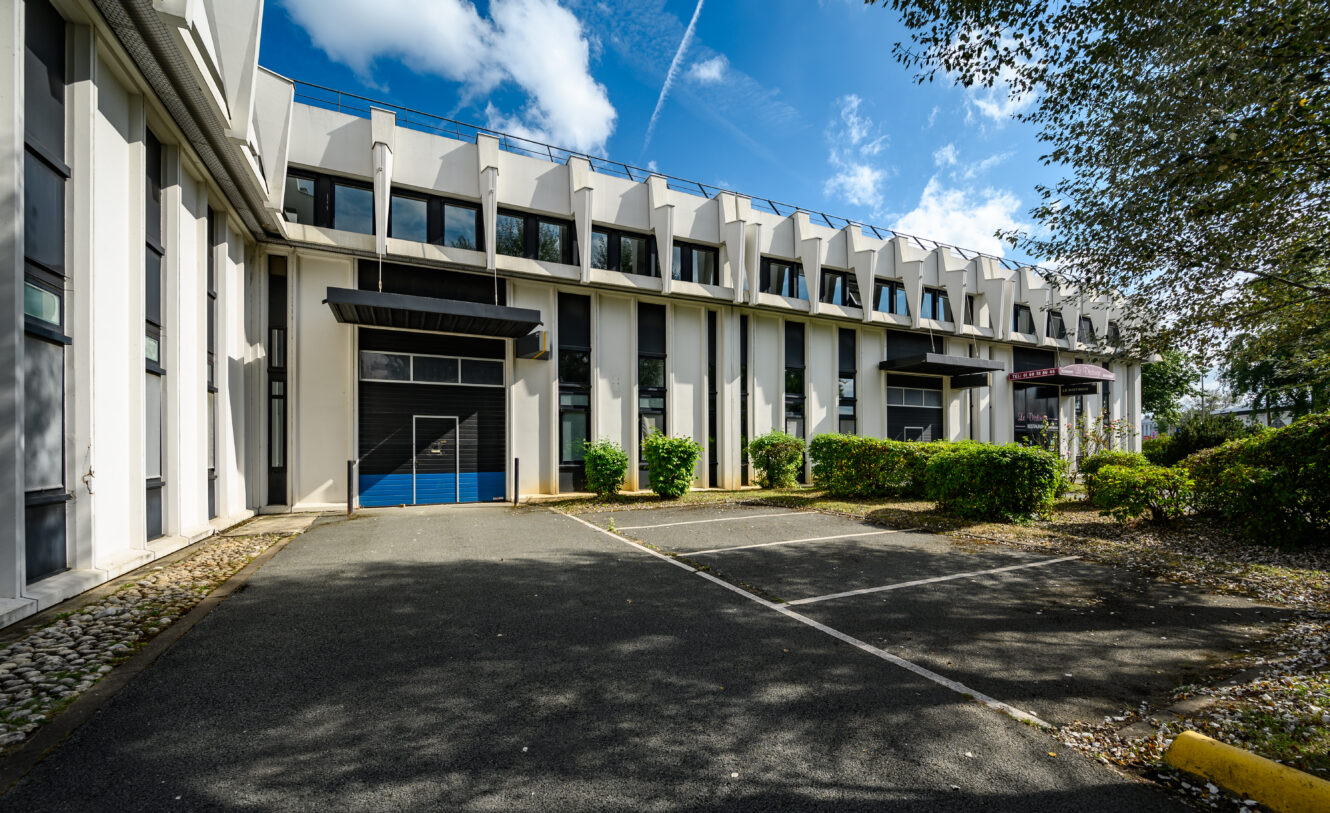 Immeuble de bureaux moderne de deux étages avec de grandes fenêtres, des colonnes verticales et un parking vide au premier plan par une journée ensoleillée.