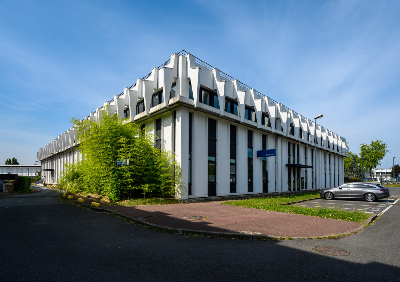 Immeuble de bureaux moderne de deux étages avec une façade géométrique blanche, de grandes fenêtres verticales et un parking adjacent sous un ciel bleu clair.
