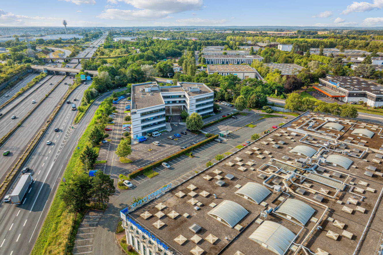 Vue aérienne d'immeubles de bureaux et d'une usine près d'une grande route avec plusieurs voitures et camions, entourés de verdure et d'arbres sous un ciel partiellement nuageux.