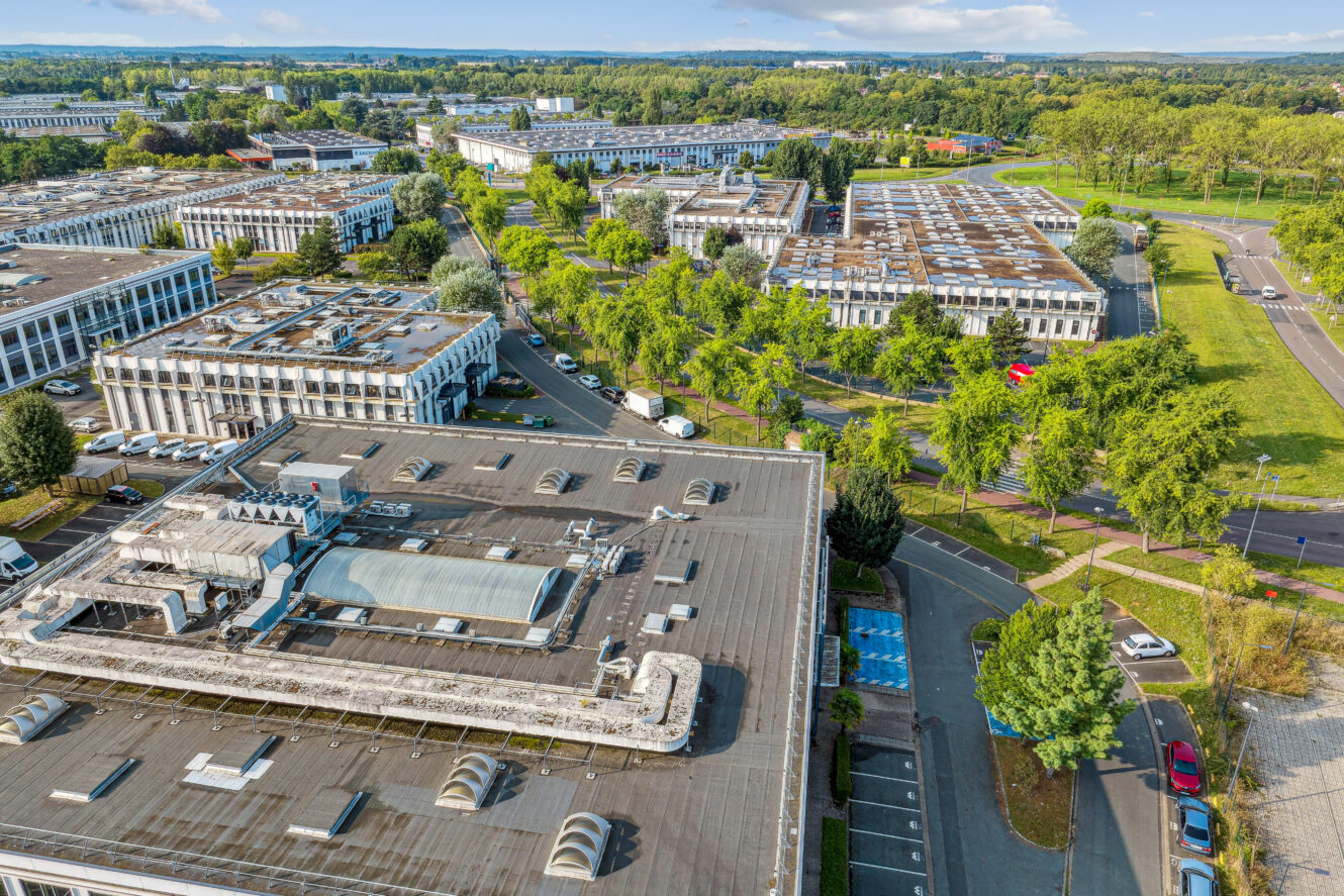 Vue aérienne d'un grand complexe industriel ou de bureaux avec de nombreux bâtiments blancs, des parkings et des arbres verts entourant la zone par temps clair.