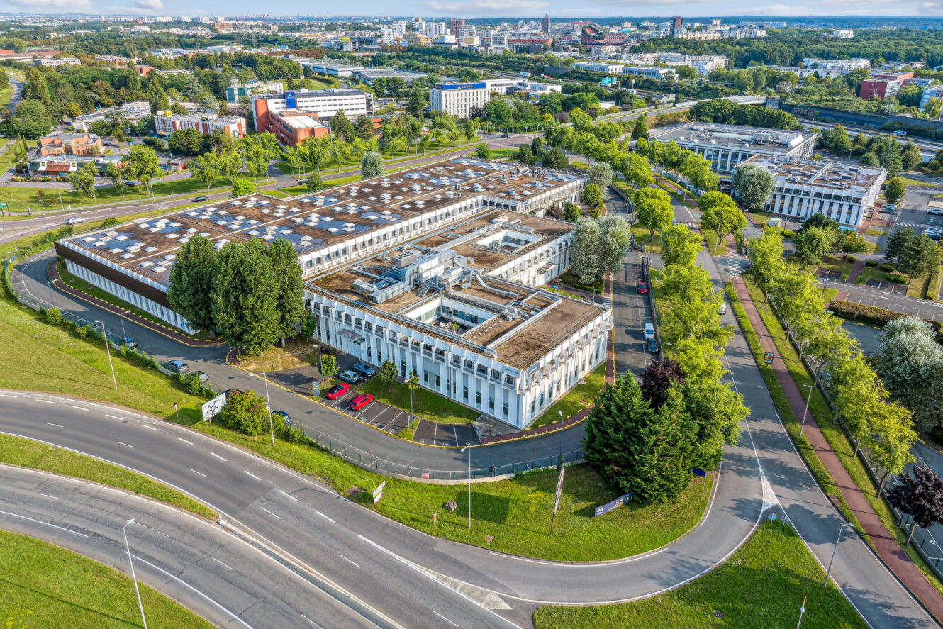 Vue aérienne d'un grand bâtiment industriel ou de bureaux rectangulaire entouré de routes, d'arbres et d'autres bâtiments dans une zone suburbaine.