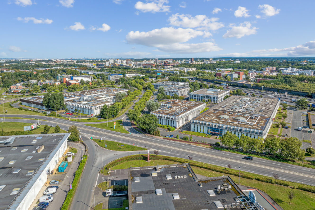 Vue aérienne d'un complexe de grands immeubles de bureaux ou industriels à toit plat, entourés de routes, d'arbres et d'un paysage urbain en arrière-plan, sous un ciel partiellement nuageux.