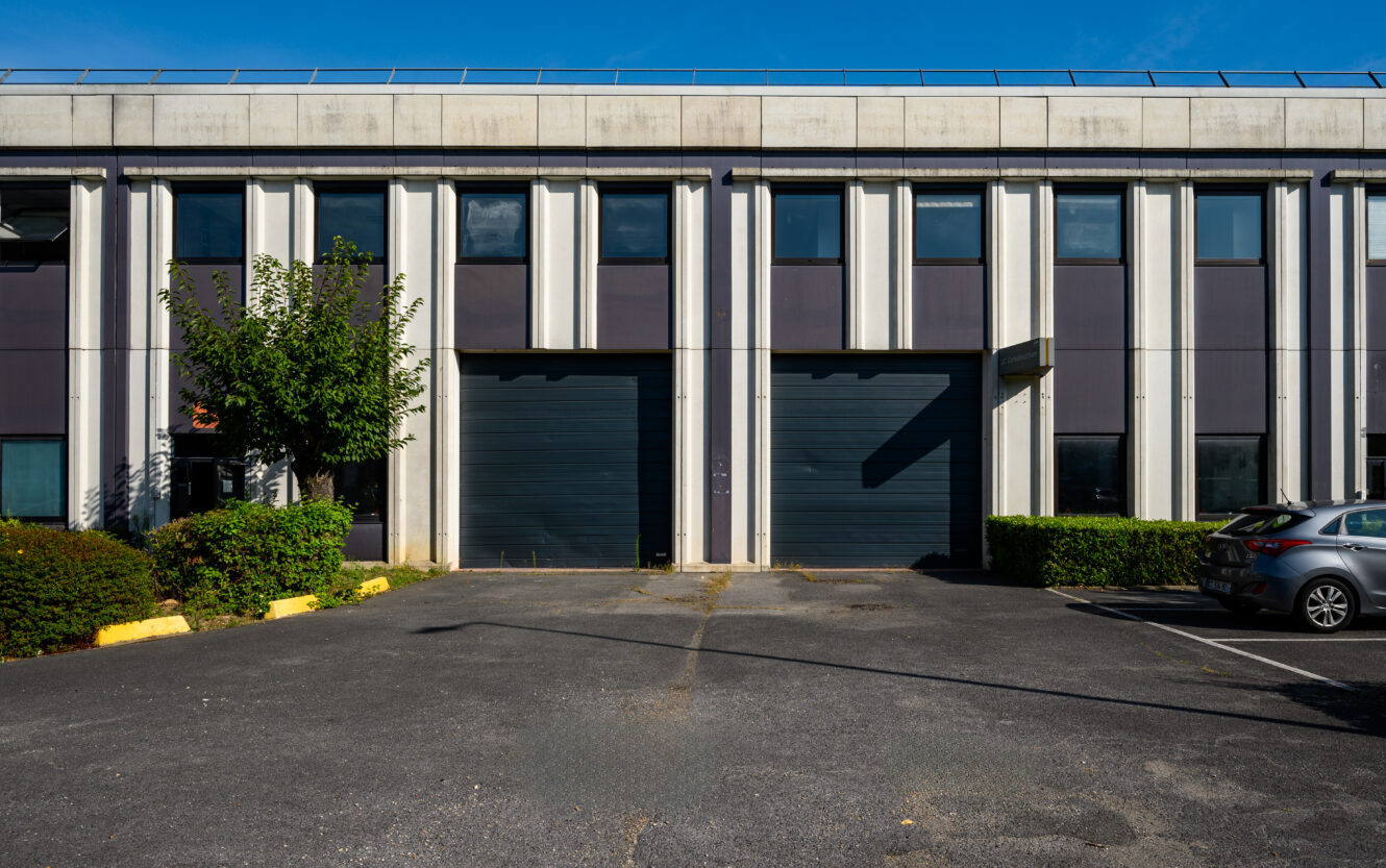 Façade d'un bâtiment industriel avec deux grandes portes de garage noires, un arbre à gauche, des haies et des voitures garées à droite dans un parking ensoleillé.