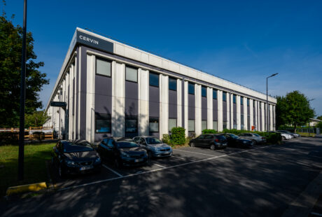 Un immeuble de bureaux de deux étages portant le nom de CERVIN, avec plusieurs voitures garées devant, entouré d'arbres et de verdure par une journée ensoleillée.