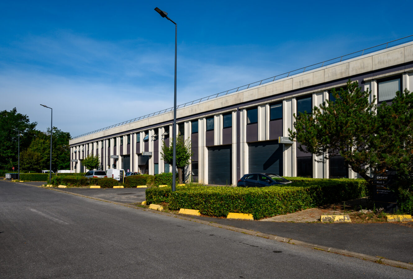 Un bâtiment commercial moderne de deux étages avec de grandes fenêtres, entouré de haies bien taillées, d'arbres et d'un parking avec plusieurs voitures garées sous un ciel bleu clair.