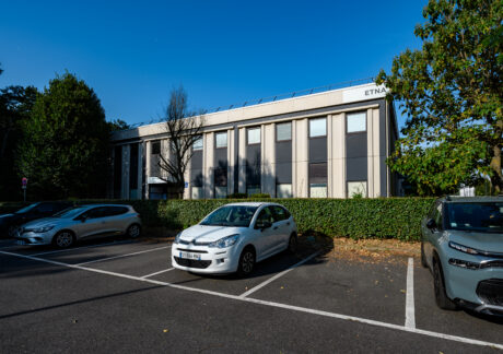 Par temps clair, une voiture blanche est garée dans un parking presque vide devant un immeuble de bureaux de deux étages portant le nom d'ETNA.
