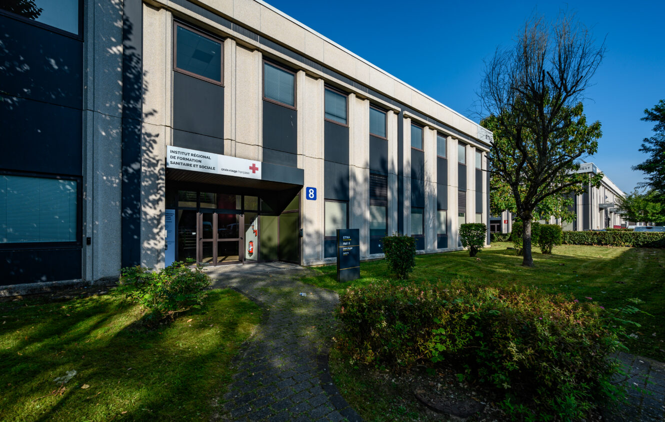 Un bâtiment moderne de deux étages avec de grandes fenêtres, portant le nom d'Institut régional de Santé et de Service Social, entouré d'herbe verte, d'arbres et d'arbustes lors d'une journée ensoleillée.
