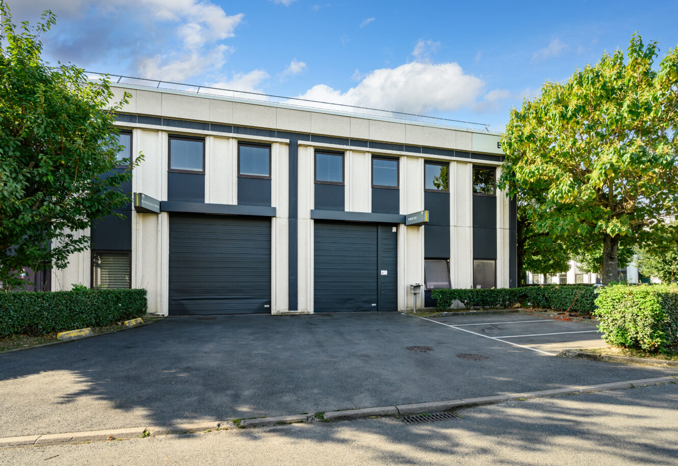 Bâtiment industriel moderne de deux étages avec deux grandes portes de garage noires, plusieurs fenêtres et des places de parking adjacentes, entouré de verdure sous un ciel partiellement nuageux.