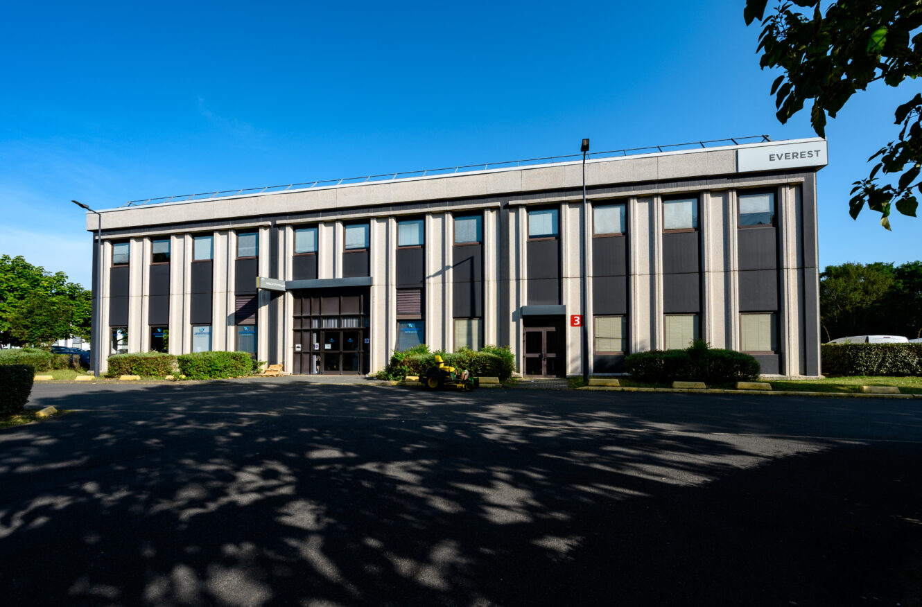 Un bâtiment commercial de deux étages avec un toit plat, appelé Everest, doté de grandes fenêtres verticales et entouré de verdure sous un ciel bleu clair.