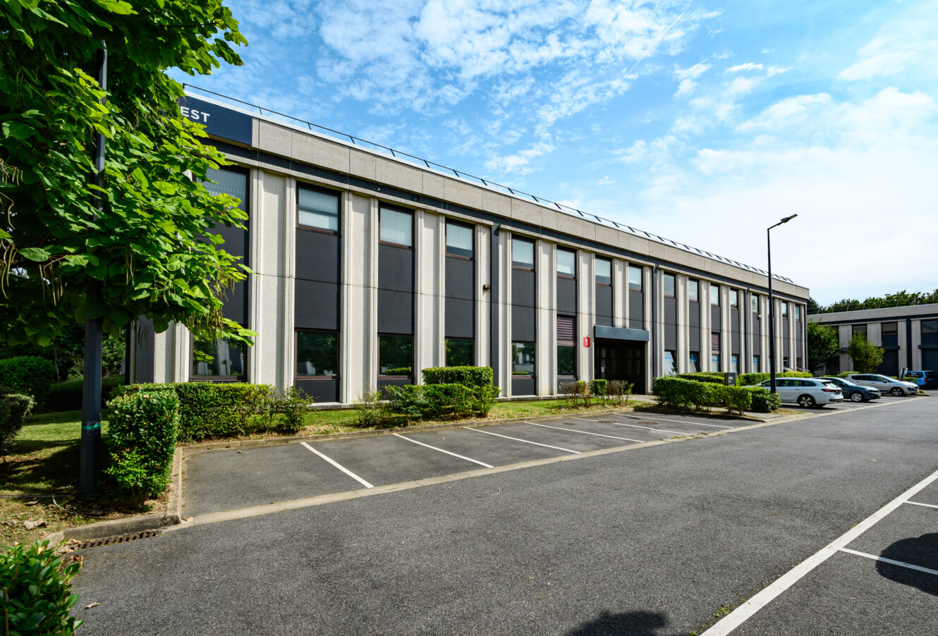 Un immeuble de bureaux moderne de deux étages avec des panneaux de fenêtres verticaux, entouré de verdure et d'un parking adjacent avec plusieurs voitures garées.
