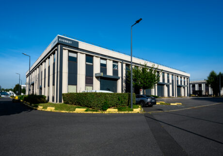Immeuble de bureaux de deux étages étiqueté EVEREST avec des fenêtres verticales, des places de parking, des haies et des arbres, situé lors d'une journée ensoleillée avec un ciel bleu clair.