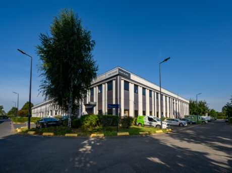 Un immeuble de bureaux de deux étages avec de grandes fenêtres, entouré de voitures garées et d'arbres, sous un ciel bleu clair.