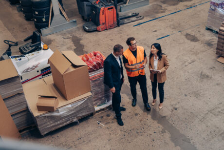 Trois personnes discutent dans un entrepôt ; l'une porte un gilet de sécurité, les deux autres sont en tenue de travail et sont entourées de palettes, de cartons et de matériaux d'emballage.