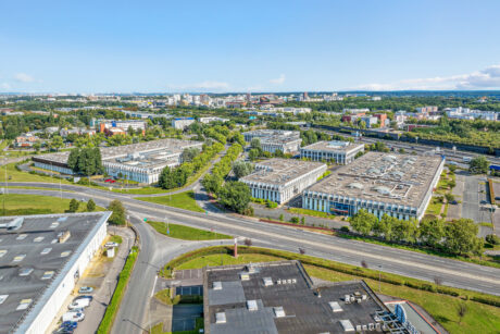 Vue aérienne d'un parc d'activités comprenant plusieurs immeubles de bureaux, des espaces verts, des routes et la zone urbaine environnante sous un ciel bleu clair.