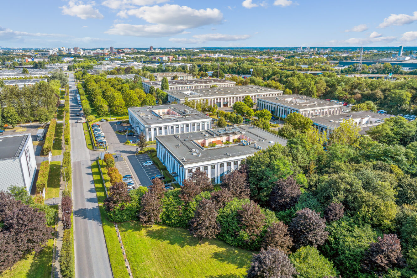 Vue aérienne d'un complexe de bureaux composé de plusieurs bâtiments blancs entourés d'arbres et de verdure, le long d'une route, sous un ciel dégagé.