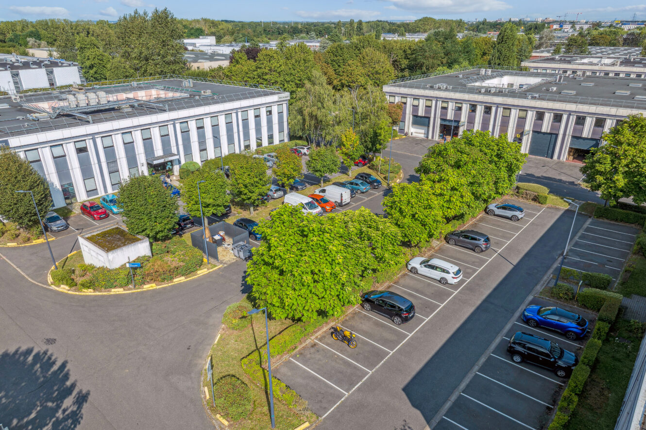 Vue aérienne d'un parking avec des voitures garées sur des places marquées, entouré d'immeubles de bureaux et d'arbres verts par une journée ensoleillée.