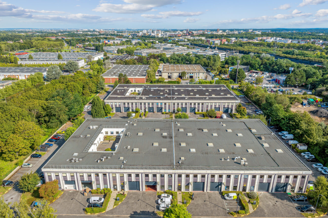 Vue aérienne de plusieurs grands immeubles de bureaux à toit plat, entourés d'arbres et de parkings, dans une zone urbaine sous un ciel partiellement nuageux.