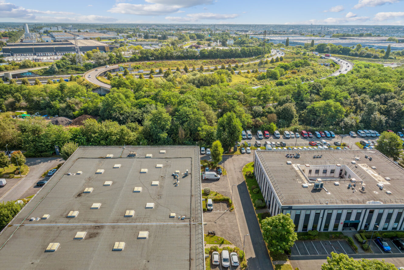 Vue aérienne de deux immeubles de bureaux avec des parkings entourés d'arbres, avec une autoroute et une zone industrielle en arrière-plan.