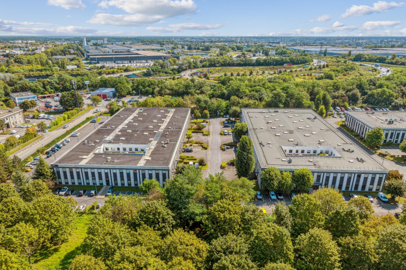 Vue aérienne de deux immeubles de bureaux rectangulaires entourés d'arbres, de parkings et de routes, avec une zone industrielle et un paysage urbain en arrière-plan.