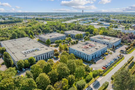 Vue aérienne de plusieurs immeubles de bureaux rectangulaires entourés d'arbres, de voitures garées et de routes, avec un paysage urbain et des espaces verts en arrière-plan, sous un ciel partiellement nuageux.