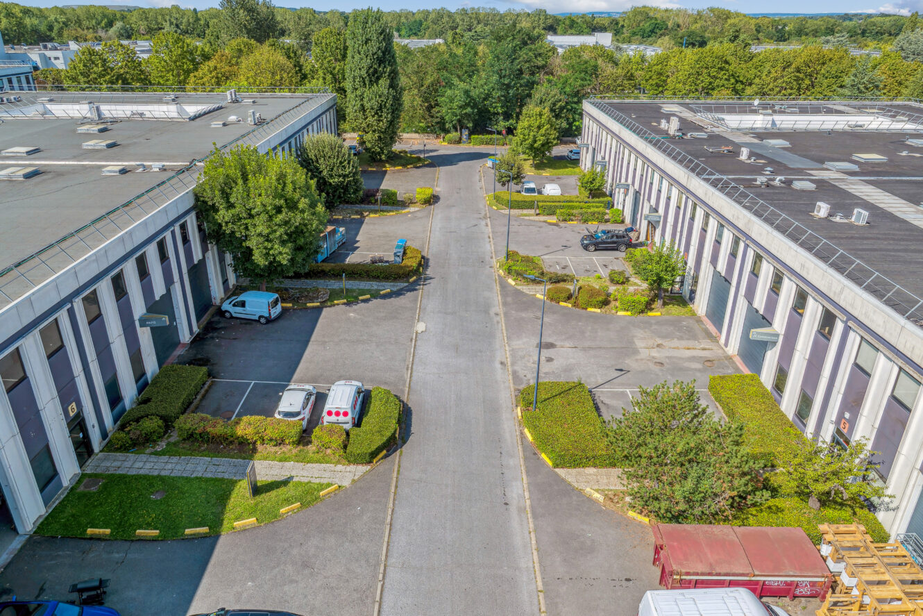 Vue aérienne d'un complexe industriel comprenant deux grands entrepôts, des véhicules en stationnement, de la verdure et une route centrale pavée.