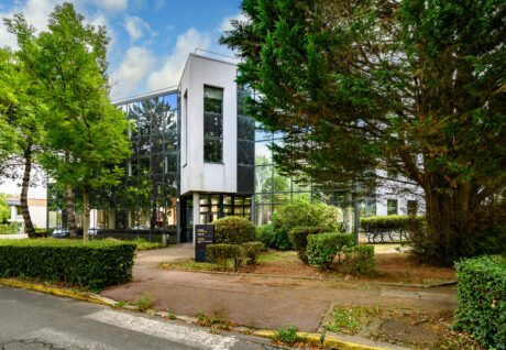 Immeuble de bureaux moderne de deux étages avec de grandes baies vitrées, entouré d'arbres, de haies et d'arbustes, par temps nuageux.