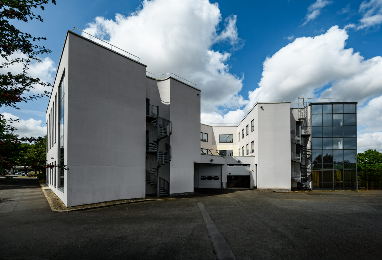 Un bâtiment moderne blanc avec des escaliers extérieurs en métal, de grandes fenêtres et une cage d'ascenseur en verre, sous un ciel partiellement nuageux.