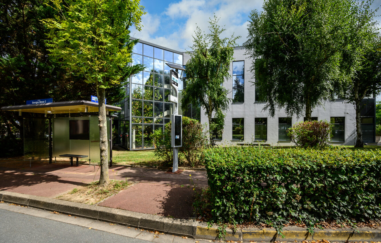 Un immeuble de bureaux en verre et en pierre blanche se dresse derrière un arrêt de bus dans une rue tranquille, entouré d'arbres et de verdure sous un ciel partiellement nuageux.