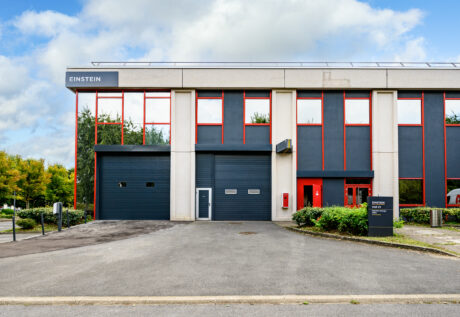 Bâtiment industriel ou de bureaux moderne avec de grandes fenêtres, des bordures rouges et un panneau indiquant Einstein au-dessus d'une porte de garage et d'une entrée sombres.