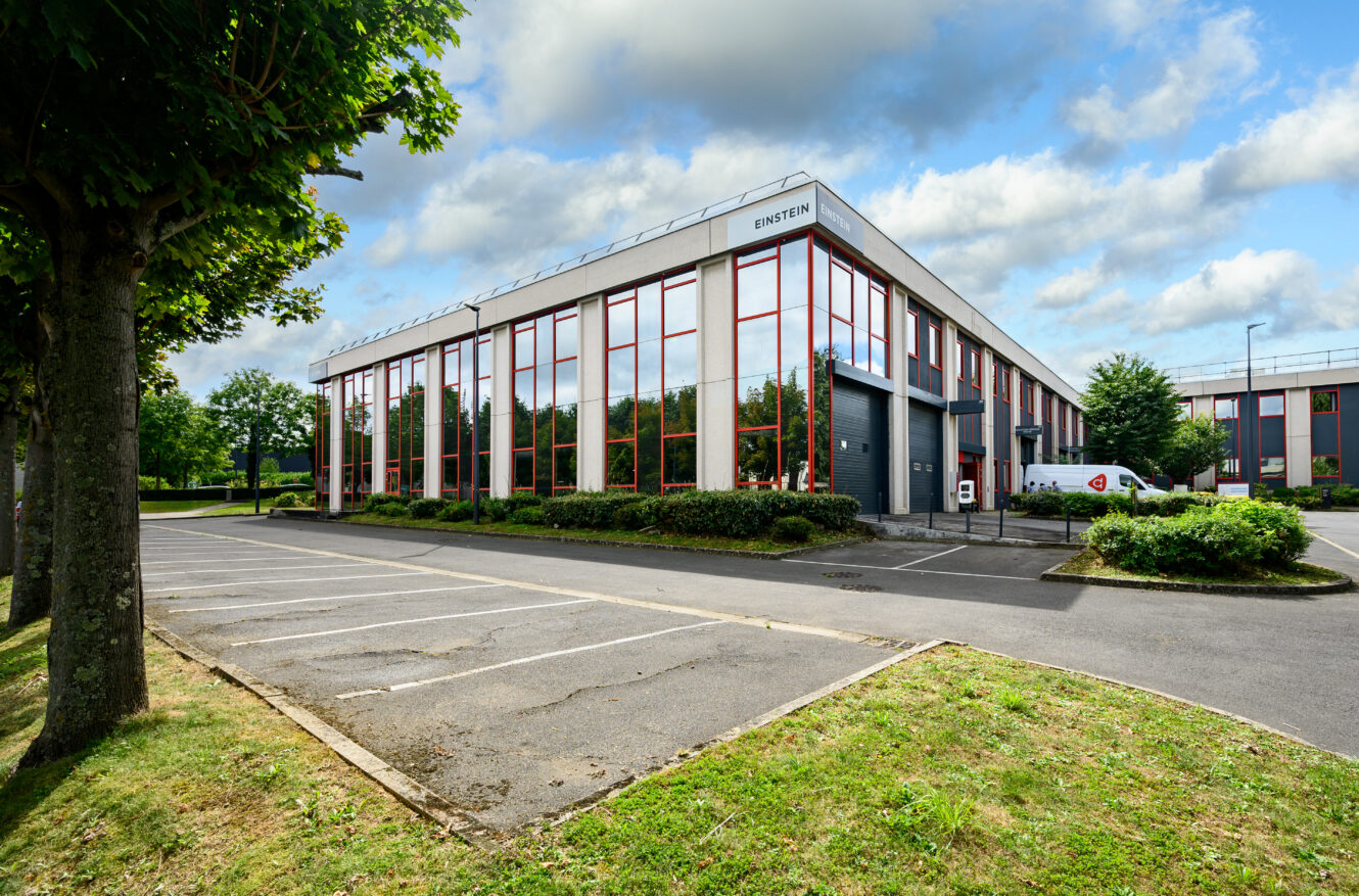 Un immeuble de bureaux moderne de deux étages étiqueté Einstein avec de grandes fenêtres, entouré d'arbres, d'un parking vide et d'un ciel partiellement nuageux.