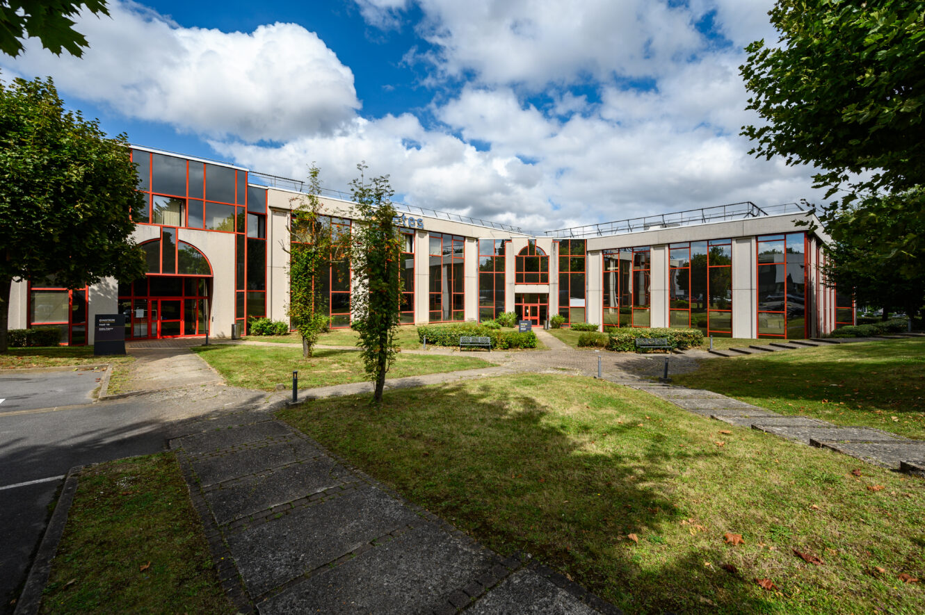 Immeuble de bureaux moderne de deux étages avec de grandes fenêtres, entouré d'herbe, de chemins pavés et d'arbres sous un ciel partiellement nuageux.