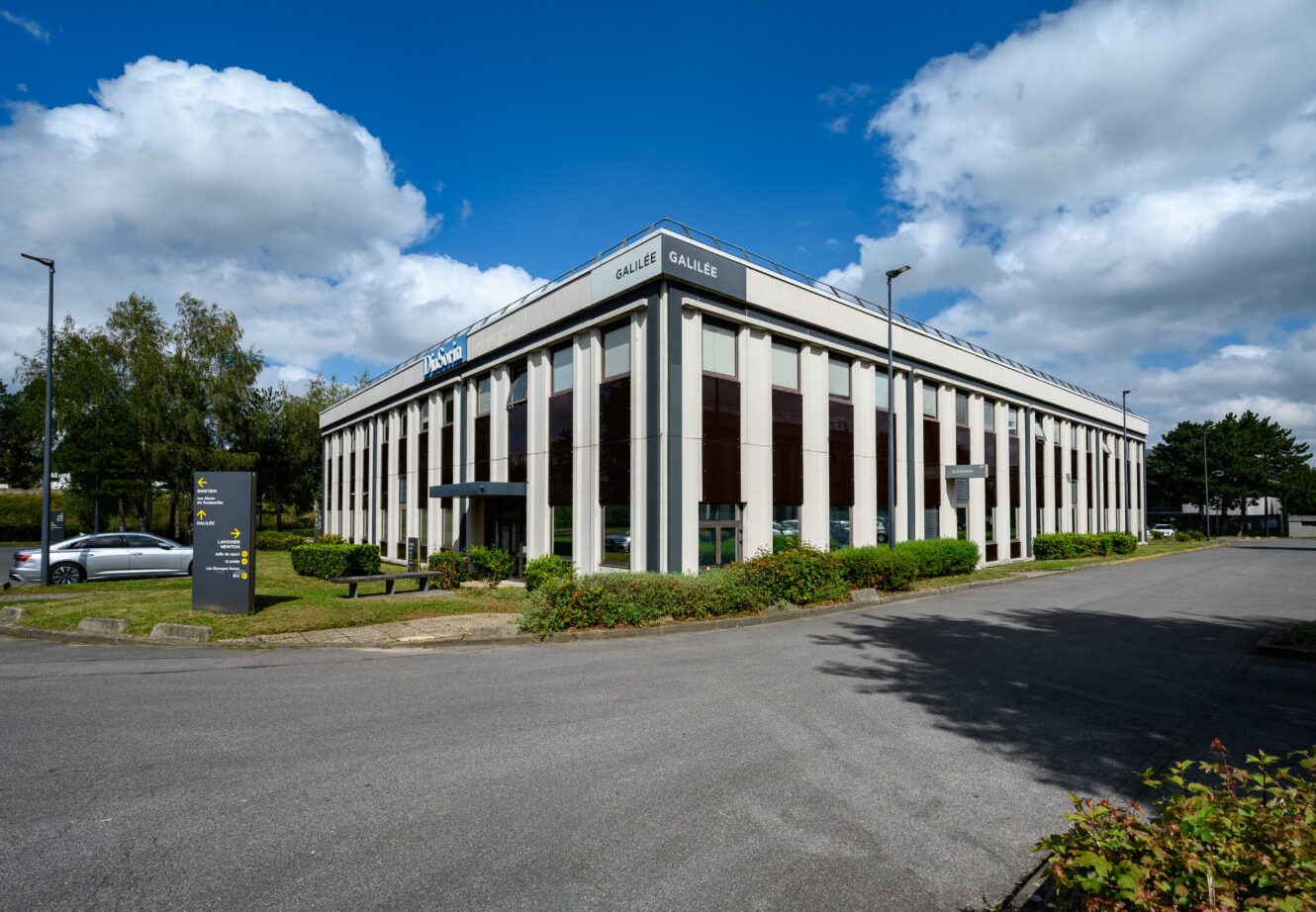 Un immeuble de bureaux de deux étages avec de grandes fenêtres et un panneau de signalisation à l'avant, entouré de verdure et de voitures garées sous un ciel partiellement nuageux.