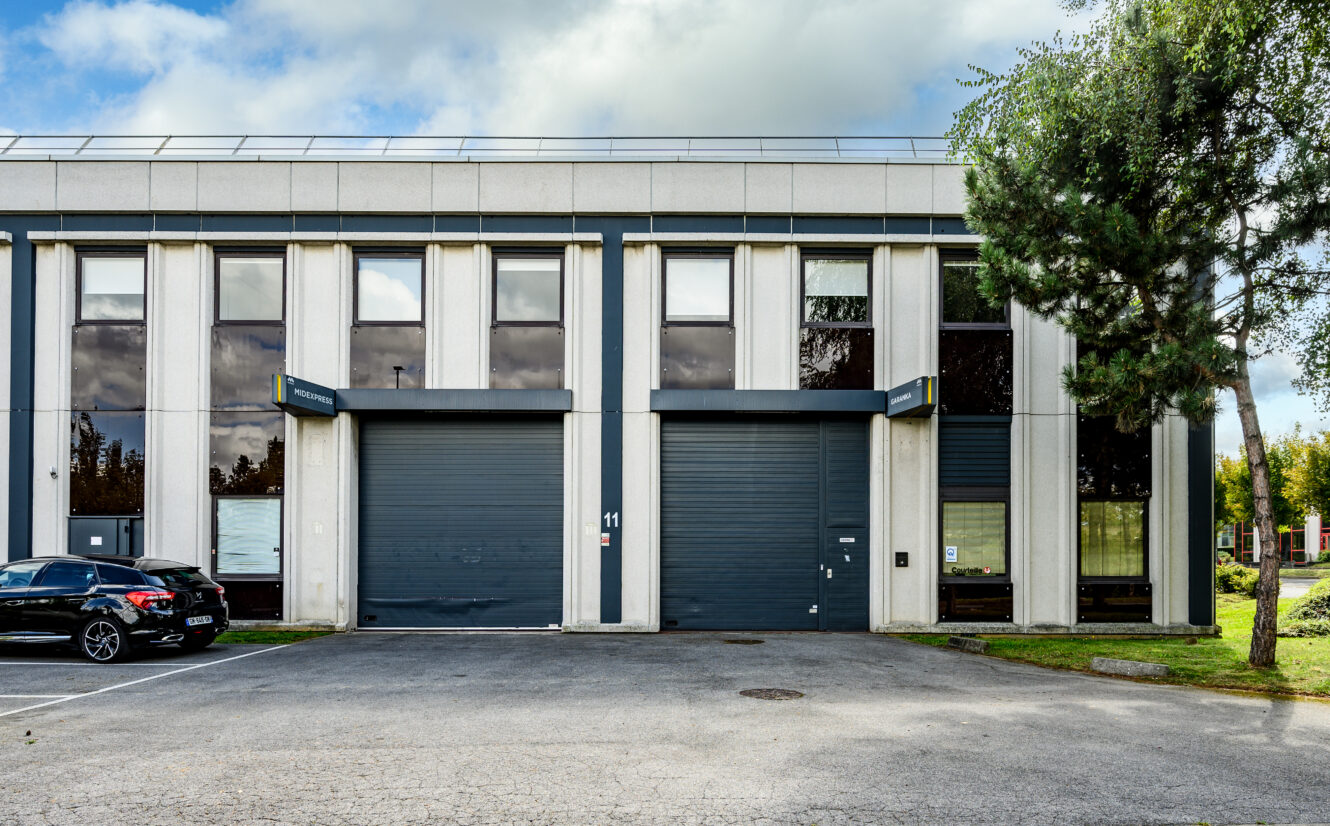 Un bâtiment industriel moderne avec de grandes baies vitrées, deux portes de garage noires, une voiture noire garée et un arbre devant.