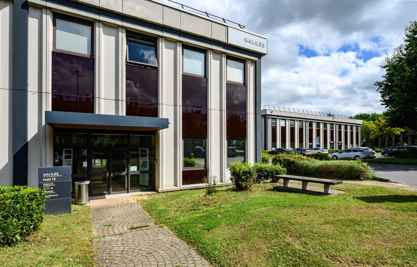 Immeuble de bureaux moderne étiqueté Galilée avec de grandes fenêtres, une enseigne près de l'entrée, un banc sur la pelouse et des voitures garées en arrière-plan sous un ciel partiellement nuageux.