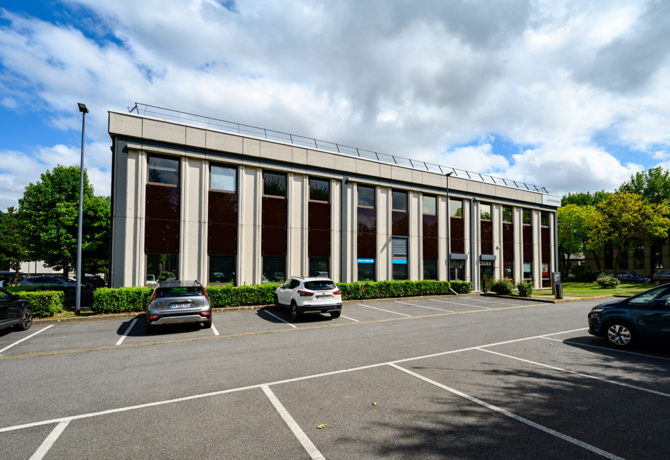 Immeuble de bureaux de deux étages avec de grandes fenêtres et plusieurs voitures garées dans le parking par une journée partiellement nuageuse.
