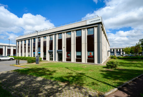Un immeuble de bureaux moderne de deux étages étiqueté Lavoisier avec de grandes fenêtres, des voitures garées à proximité et un espace herbeux devant sous un ciel partiellement nuageux.