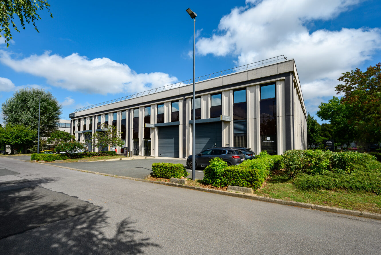 Un immeuble de bureaux moderne de deux étages avec de grandes fenêtres, un petit parking avec plusieurs voitures, de la verdure et un ciel bleu clair avec des nuages épars.