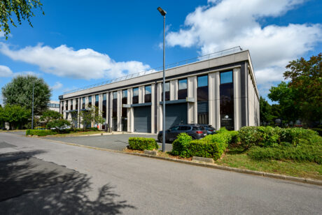 Un immeuble de bureaux moderne de deux étages avec de grandes fenêtres, un petit parking avec plusieurs voitures, de la verdure et un ciel bleu clair avec des nuages épars.