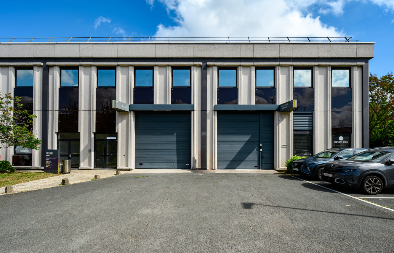 Bâtiment commercial moderne avec de grandes fenêtres, deux portes grises à enroulement et des voitures garées devant, sous un ciel partiellement nuageux.