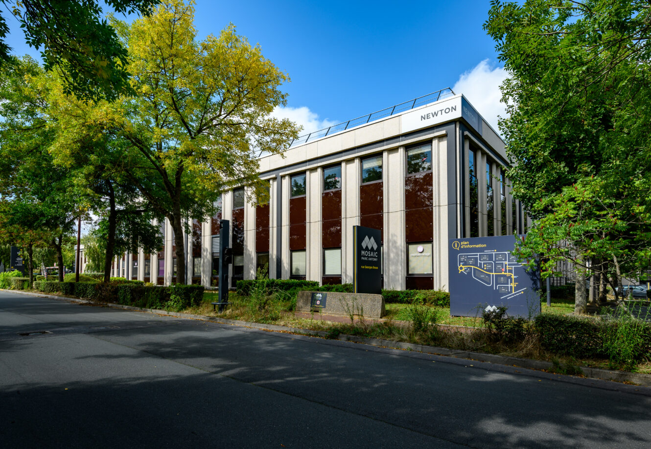 Un immeuble de bureaux moderne de deux étages, étiqueté NEWTON, avec de grandes fenêtres, entouré d'arbres, avec des panneaux de signalisation pour Mosaic et un plan du campus visible près de l'entrée.