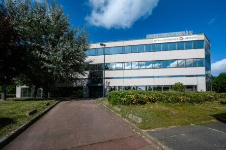 Un immeuble de bureaux de trois étages avec une enseigne Stallergenes Greer, des fenêtres réfléchissantes et de la verdure environnante sous un ciel partiellement nuageux.