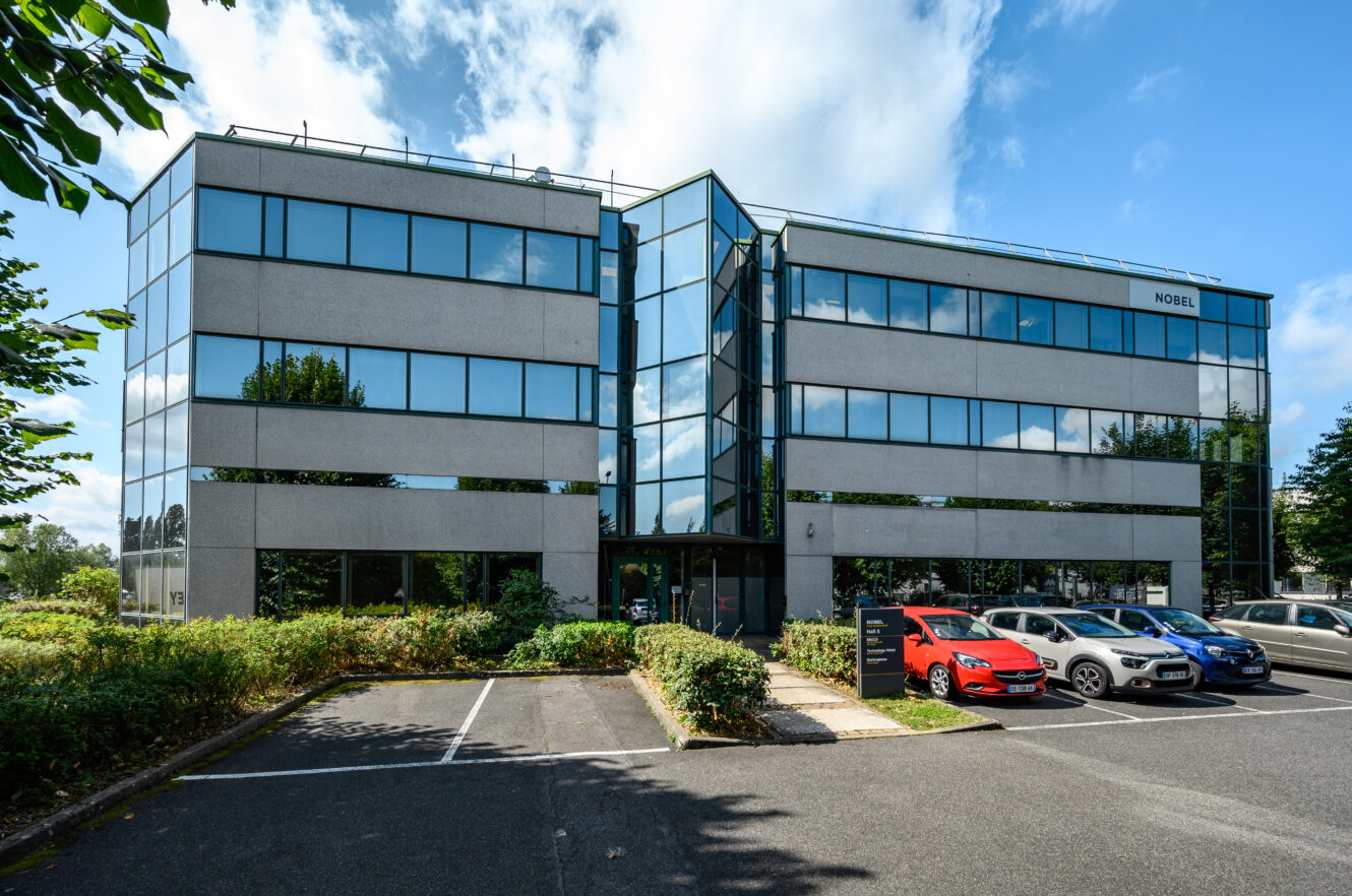 Immeuble de bureaux moderne de quatre étages avec une section centrale en verre, entouré de verdure, de voitures garées et d'un parking en grande partie vide, sous un ciel partiellement nuageux.