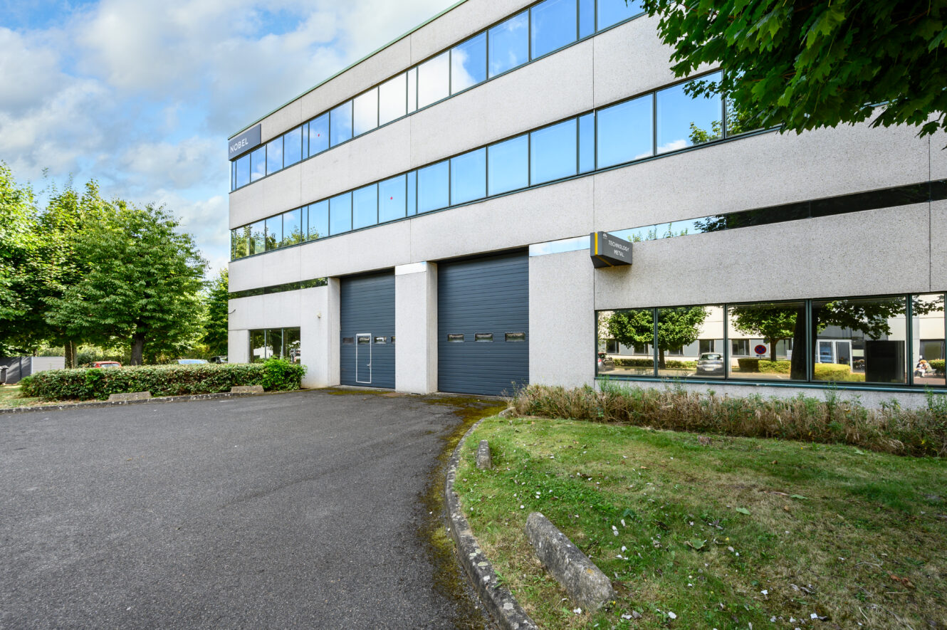Un immeuble de bureaux moderne de trois étages avec de grandes fenêtres, deux portes de garage grises et la verdure environnante par temps clair.