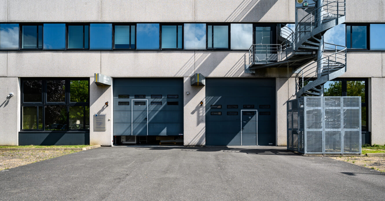 Extérieur d'un bâtiment industriel avec deux grandes portes de garage grises, des fenêtres au-dessus, un escalier extérieur en métal et une zone utilitaire clôturée sur la droite.