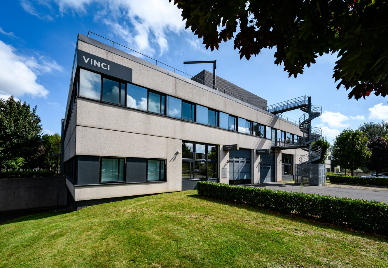 Immeuble de bureaux moderne de deux étages avec de grandes fenêtres, étiqueté VINCI sur la façade, entouré d'herbe, d'arbres et d'un escalier métallique en colimaçon sur le côté.