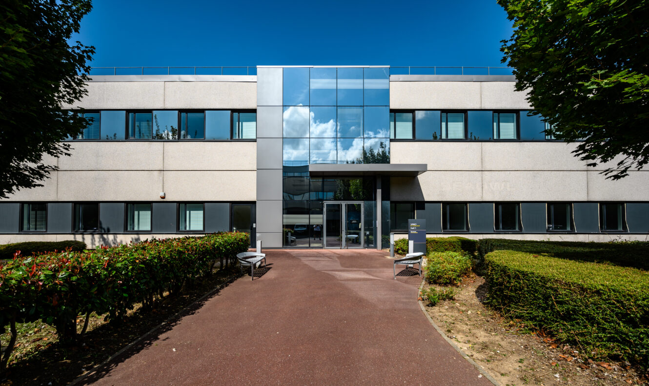Immeuble de bureaux moderne de deux étages avec une grande entrée vitrée, entouré de buissons et d'arbres paysagers sous un ciel bleu clair.