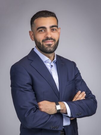 Un homme en costume bleu marine et chemise bleu clair se tient debout, les bras croisés, face à l'appareil photo, sur un fond gris uni.