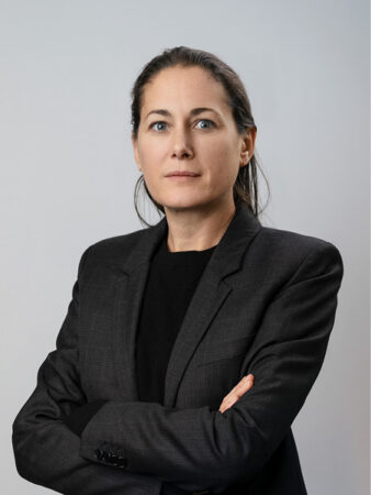 Une femme aux longs cheveux bruns, vêtue d'un blazer noir, se tient debout, les bras croisés, sur un fond gris clair uni.