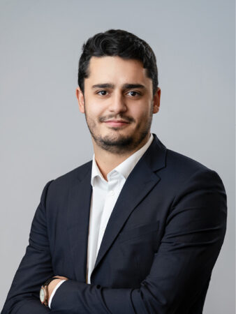 Un homme en costume sombre et chemise blanche se tient debout, les bras croisés, regardant l'appareil photo sur un fond gris clair uni.